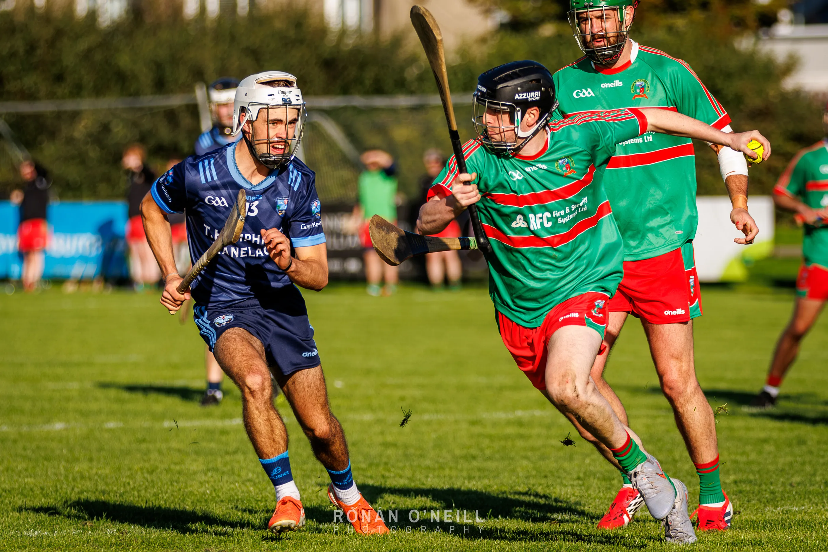 A Naomh Barróg hurler in green and red carries the sliotar upfield with a Ranelagh Gaels player in navy tracking him at Pairc Barróg.