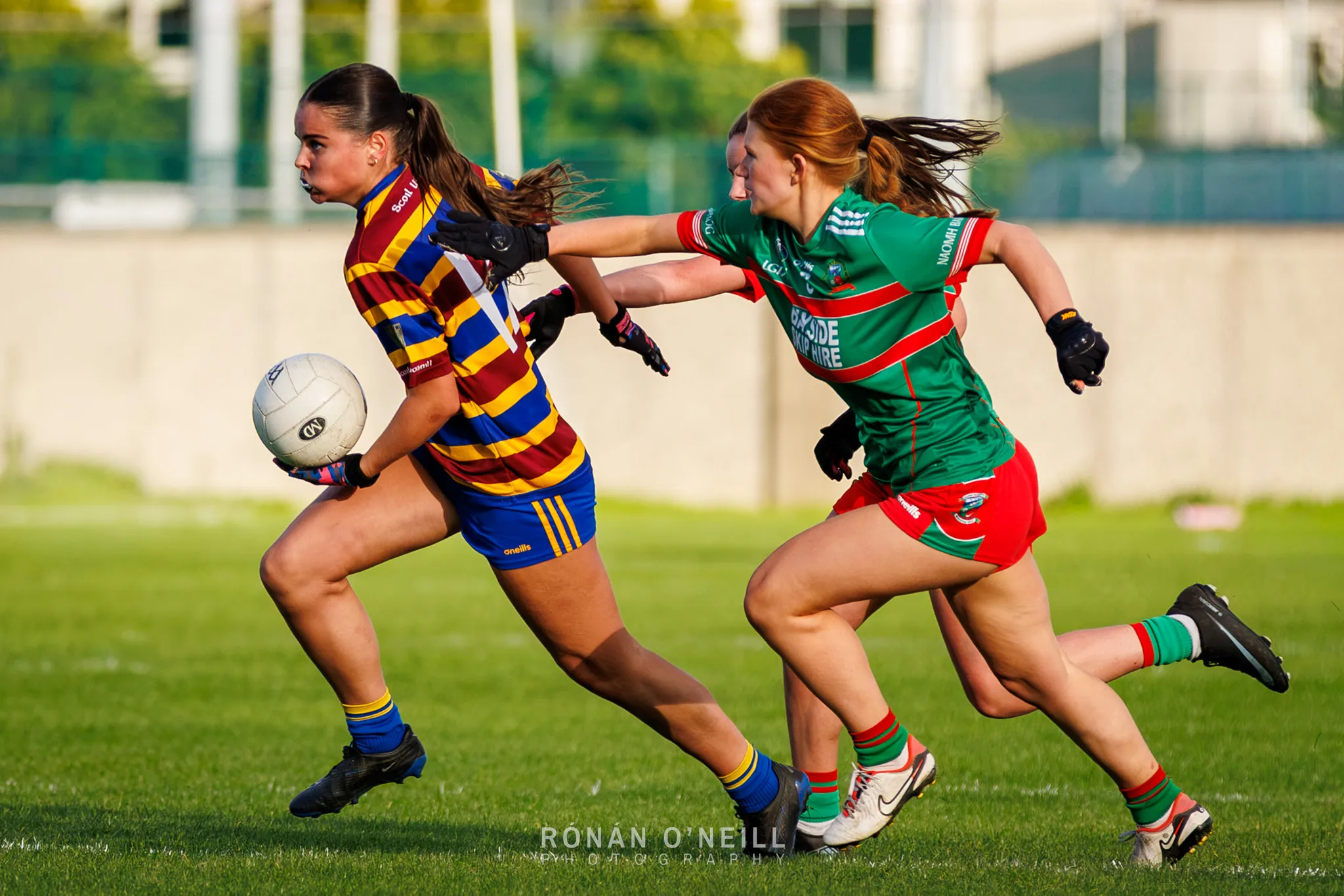 A Scoil Uí Chonaill player in red, yellow and blue hoops carries the ball upfield with two Naomh Barróg players in green and red chasing at Scoil Uí Chonaill on Clontarf Road.